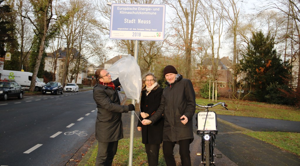 Bürgermeister Reiner Breuer (links), Umweltdezernent Dr. Matthias Welpmann und Henrike Mölleken, Leiterin des Amtes für Stadtgrün, Umwelt und Klima, enthüllen das Schild der EEA-Auszeichnung 2018 auf der Deutschen Straße.