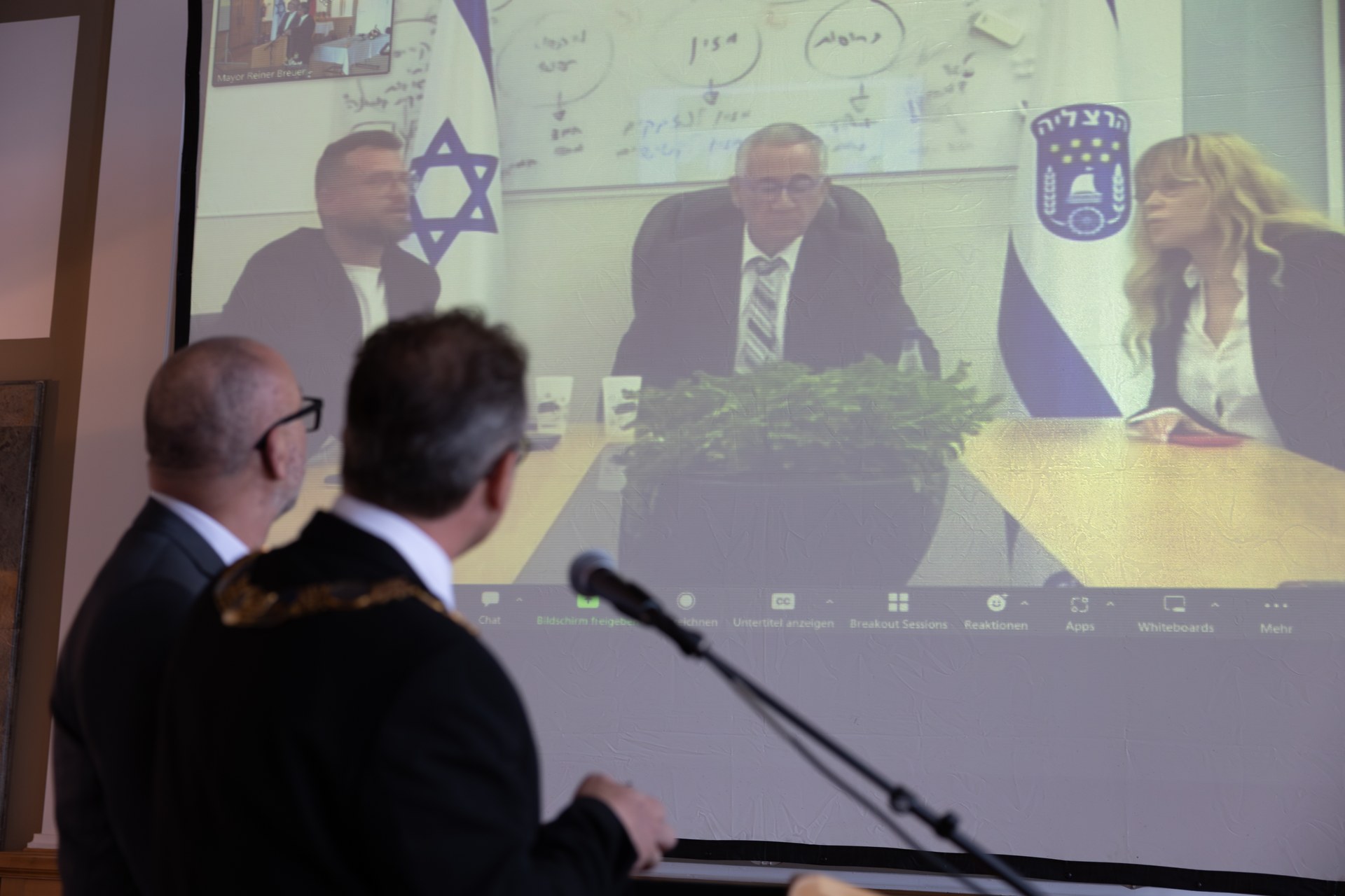 Herzliya Unterzeichnung Urkunde Städtepartnerschaft.jpg Bert Römgens und Bürgermeister Reiner Breuer stehen von einer Leinwand auf welcher der Bürgermeister aus Herzliya Moshe Fadlon sowie die stellvertretende Bürgermeisterin Ofra Bell und der Stadtdirektor Ehud Lazar zu sehen sind.