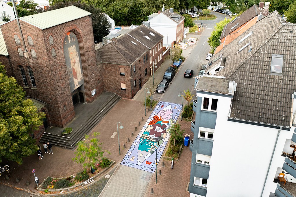 Kunstwerk im Vorbereich der Kirche St. Barbara von Konstantin Zayka Blick aus großer Höhe auf die Straße. Links ist die Kirche St. Barbara zu sehen. Das Kunstwerk auf der Straße wirkt fast wie ein farbenfroher Teppich.