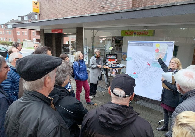 stadt-neuss_ortsmittelpunkte_stadtteilspaziergang-uedesheim-2019-10-08.jpg Eine größere Gruppe Menschen versammelt sich vor einem Geschäft. Eine Mitarbeiterin der Stadt deutet auf eine Schautafel zum Stadtspaziergang und redet.